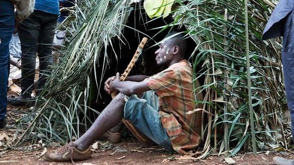 Eine Person der Neuen Auftraggeber von Kamerun sitzt zwischen grünen Zweigen auf dem Erdboden