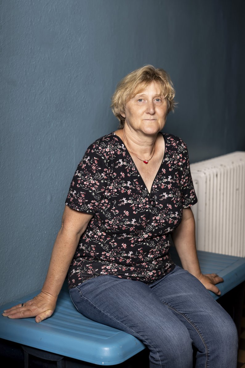 A woman with short blonde hair sits on a blue bench in front of a blue wall and looks into the camera.