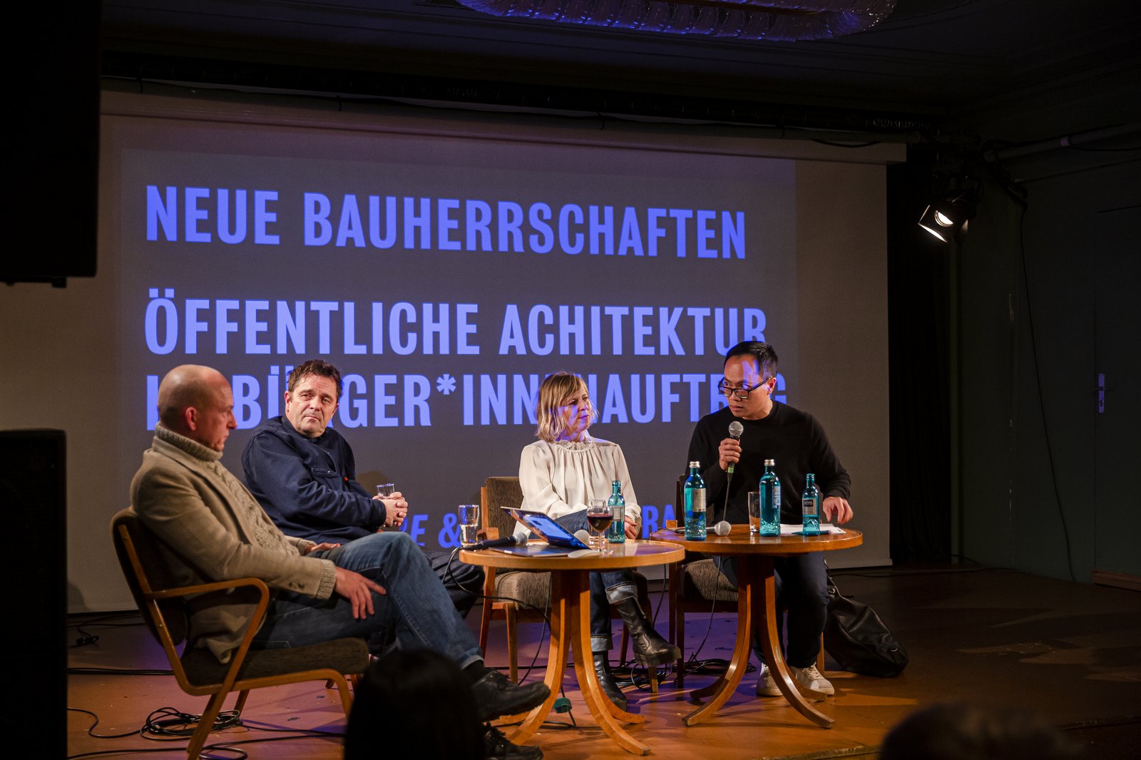 Vier Personen sitzen auf einer Bühne um einen Tisch. Eine Person spricht in ein Mikrofon. Im Hintergrund ist eine Projektion: Neue Bauherrschaften. Öffentliche Architektur im Bürger*innenauftrag.