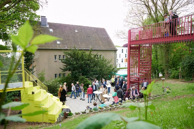 Platz in einer Grünanlage mit Menschen an Stehtischen. Links und rechts befinden sich gelbe und pinke Treppen und Brücken in die Grünanlage hinein und im Hintergrund sind ältere und neue Gebäude.