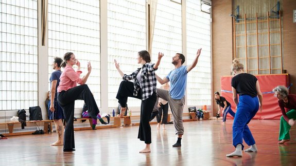Mehrere Personen der Neuen Auftraggeber tanzen in einem Saal in Marl, heben Arme und Beine in die Luft