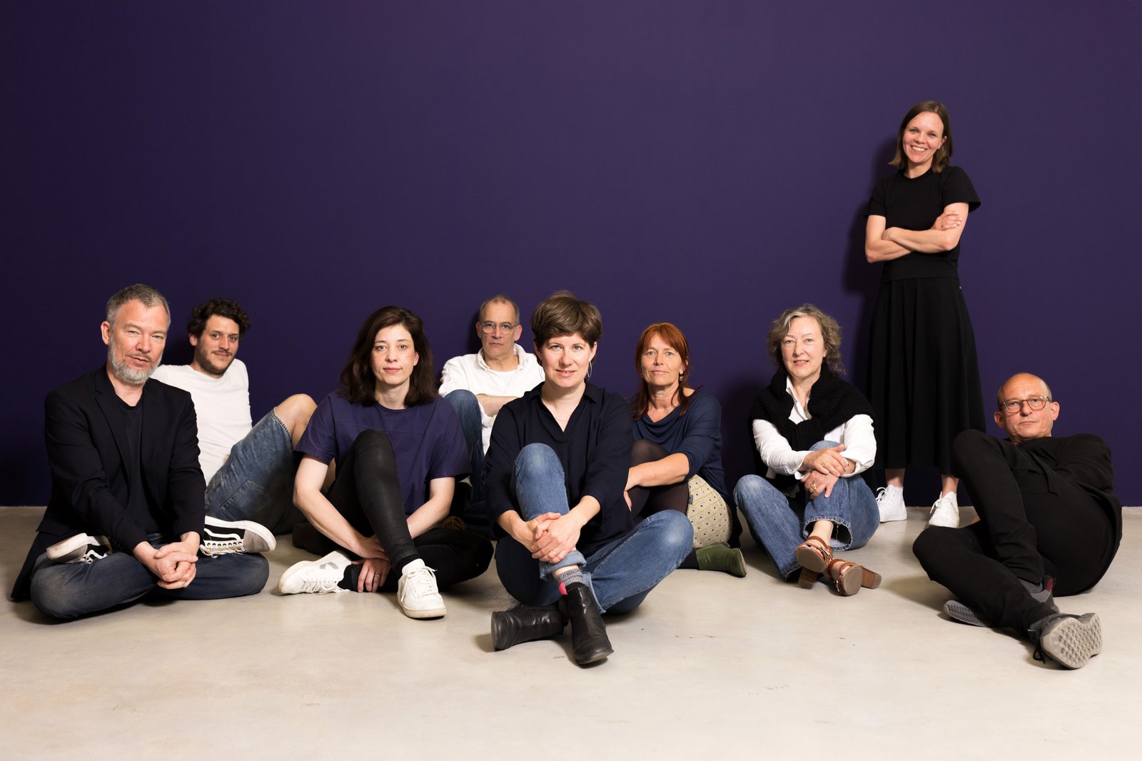 Teammitglieder der Neuen Auftraggeber stehen und sitzen vor einer blauen Wand