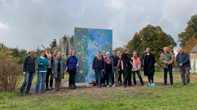 Gruppenfoto der Auftraggebergruppe vor dem Mosaik nach dem Entwurf „Tiere und Pflanzen in Wietstock" mit Künstlerin Antje Majewski und Mediatorin Susanne Burmester 