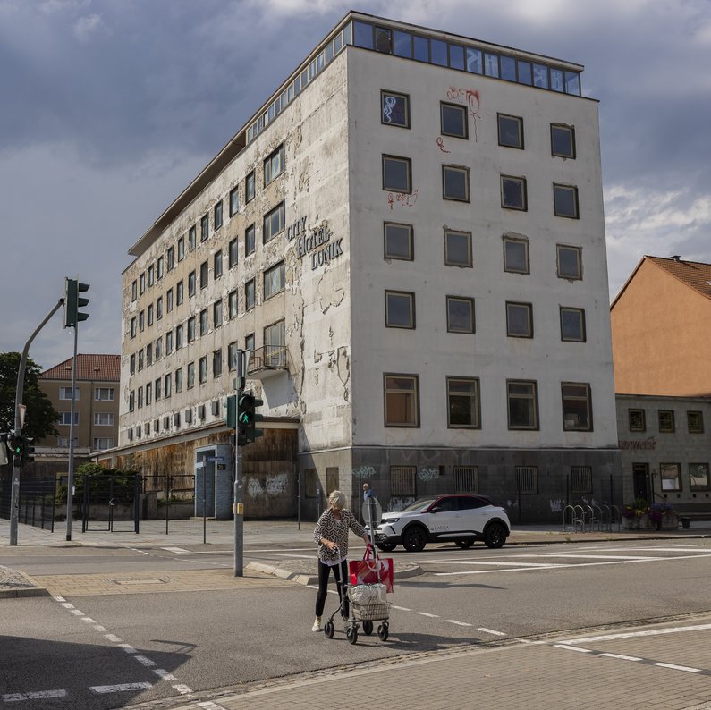 Street junction with a five-storey Plattenbau building with the lettering "City Hotel Lunik". The plaster on the façade is peeling off over a large area. A woman with a walking frame crosses the intersection in front of a red bus.