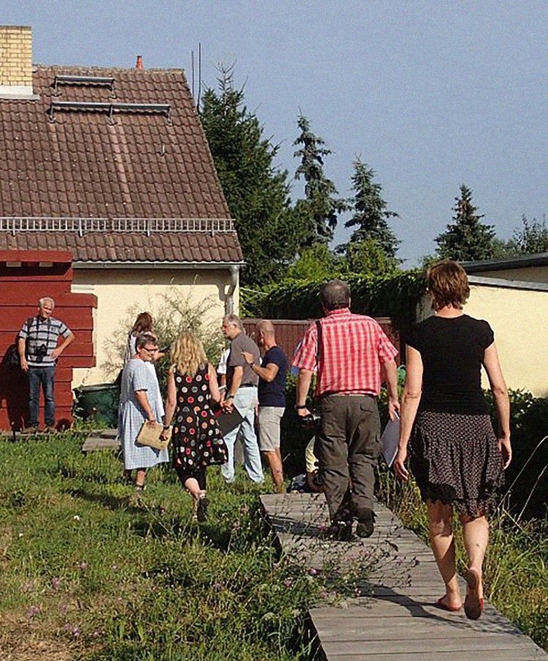 Several members of The New Patrons of Kleinliebenau walk on the wooden footbridge " Pleasure Walk " towards a house. 