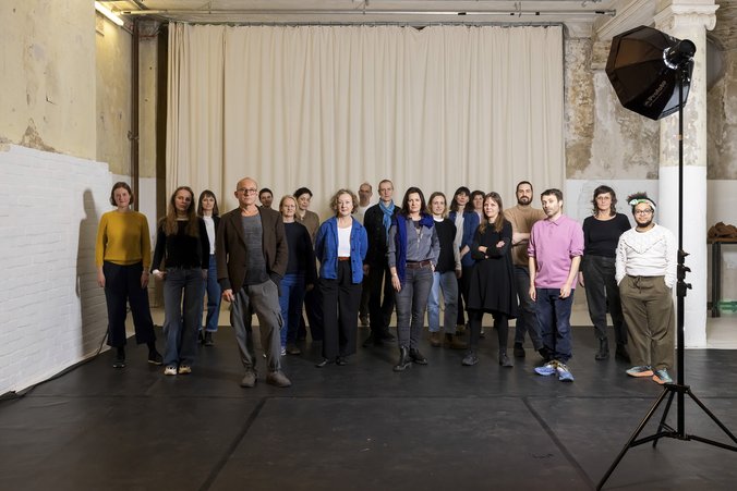 Group picture with 19 people in front of a beige curtain. There is a spotlight on a tripod in front of the group on the right.