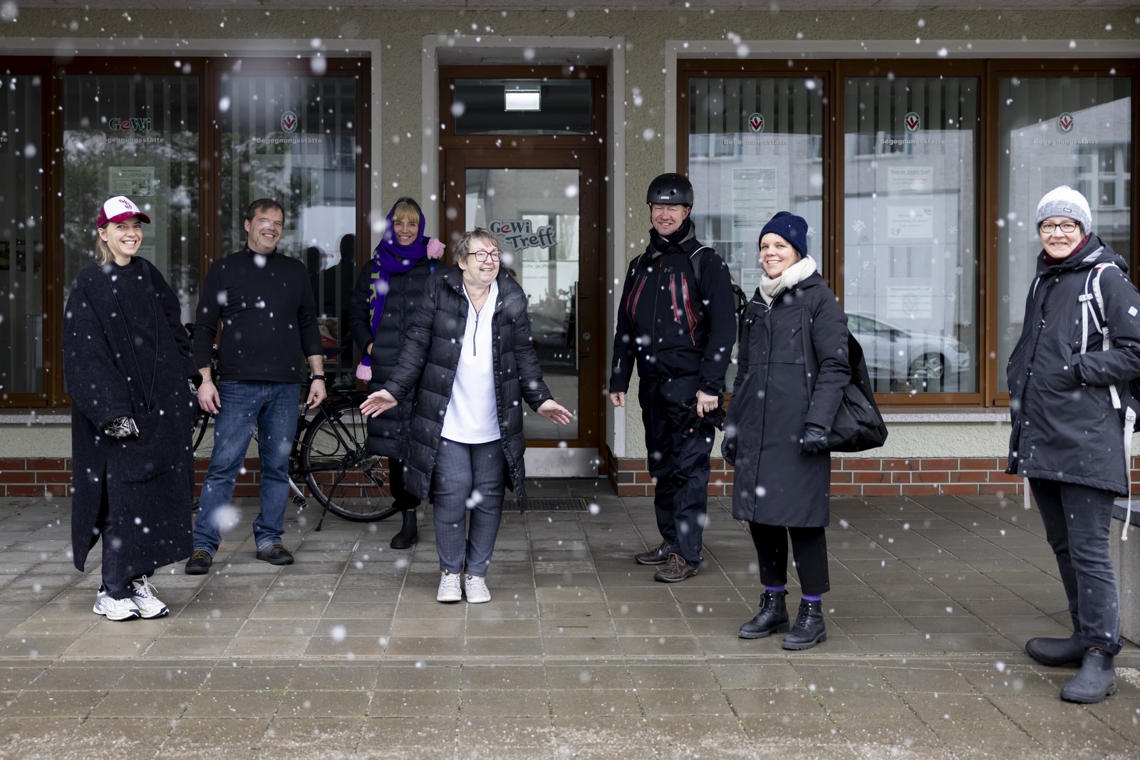 Sieben Personen stehen nebeneinander draußen vor einem Gebäude. Es schneit und sie tragen dicke Winterkleidung. Die Person in der Mitte zieht die Schultern wegen dem Schneetreiben hoch. Alle Personen lachen.