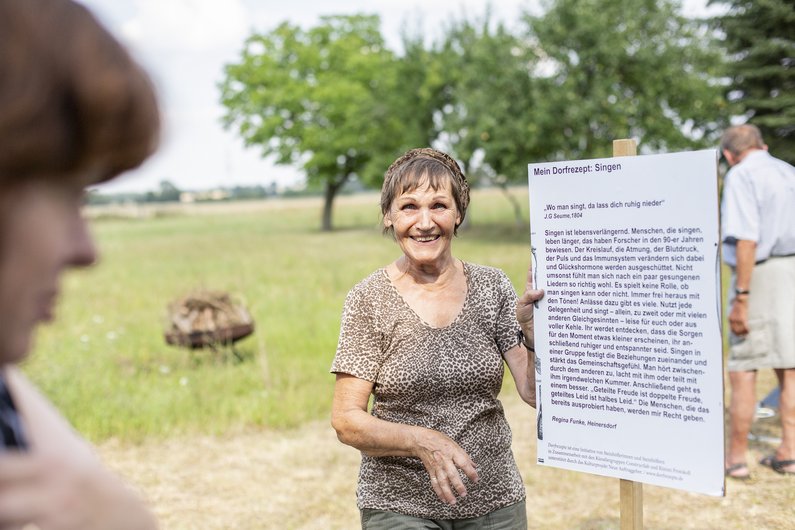 Auftraggeberin Regina Funke steht auf einem Feld in Steinhöfel und hält ein Dorfrezept in der Hand.