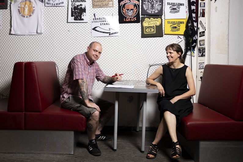 A patron in a checked shirt and the mediator sit opposite each other at a bar table. The patron is holding a pen in his hand. Punk music record covers and posters hang on the wall in the background.