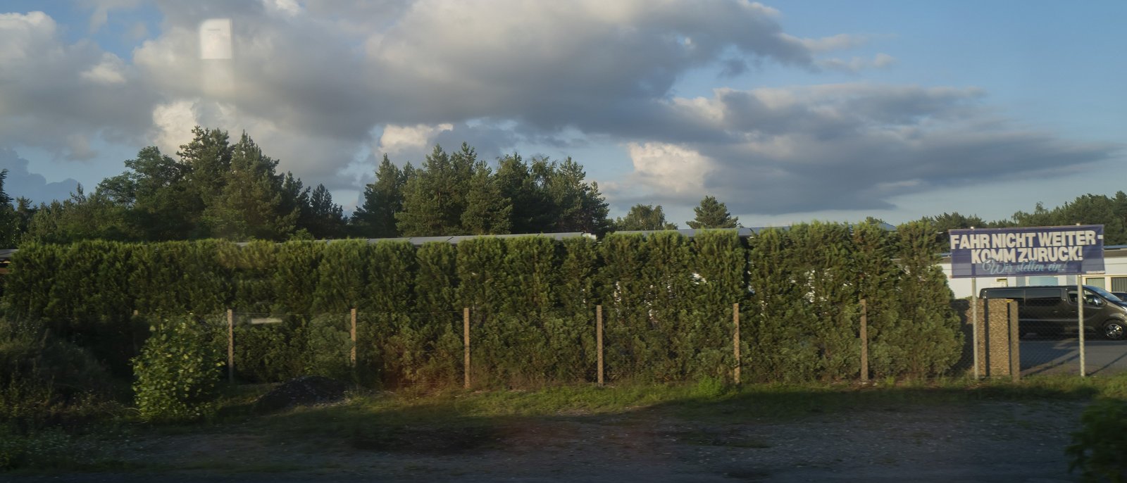 Eine grüne Gartenhecke mit einem verwitterten Schild am rechten Bildrand, auf dem steht: Fahr nicht weiter, Komm zurück.