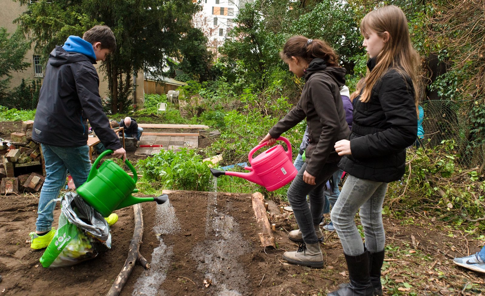 Die Neuen Auftraggeber von Mönchengladbach Drei Kinder gießen mit bunten Gießkannen ein frisch gegrabenes Beet.