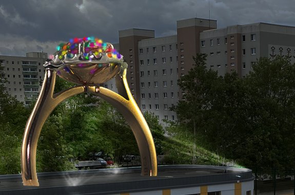 A sculpture in the shape of a golden ring with gemstones stands on the roof of a low-rise building. It is night and the gemstones of the ring send glowing rays into the dark sky. In the background are the outlines of a Plattenbau district.