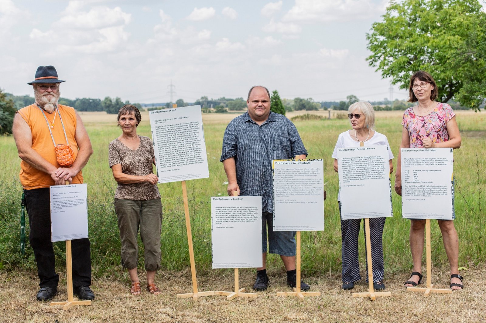 Mehrere Personen der Neuen Auftraggeber von Steinhöfel präsentieren ihre Dorfrezepte auf großen Schildern an einem Feldrand 