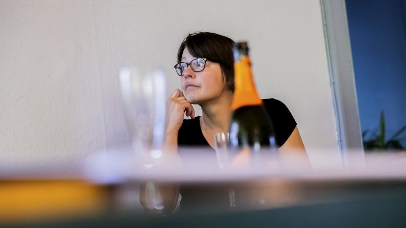 A woman with short dark hair rests her chin on her hand and looks attentively to the left. In front of her is a champagne bottle and a champagne glass. 