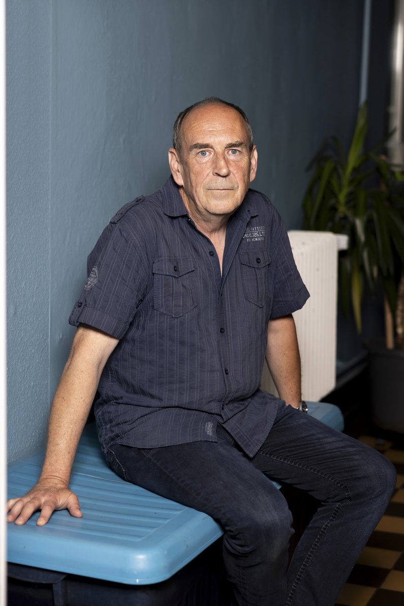 A man in a blue shirt and blue trousers sits on a blue plastic bench in front of a blue indoor wall.