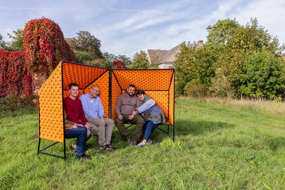 The Züsedom-Bänke are standing in a field, members of The New Patrons and mediator Holger Friese are sitting in them and laughing.