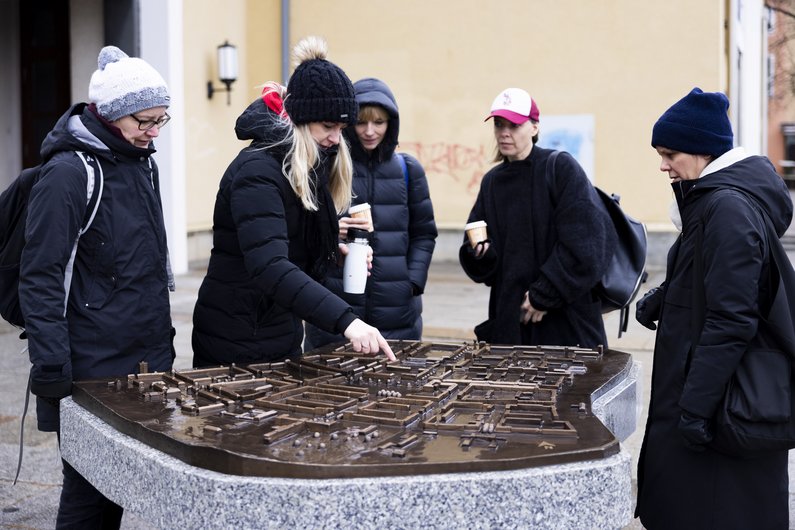 Fünf Personen in dicker Winterkleidung beugen sich über eine bronzene Miniatur-Stadtansicht. Eine Person zeigt mit dem Finger auf die Bronze und erklärt etwas.