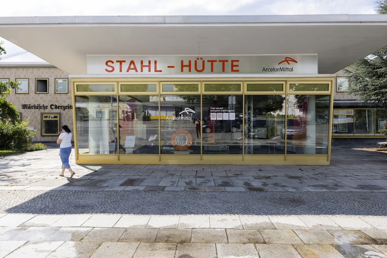 Glass pavilion with golden frame and projecting roof in front of a Plattenbau building. "Stahl-Hütte" is written in red letters above the glass front of the pavilion.