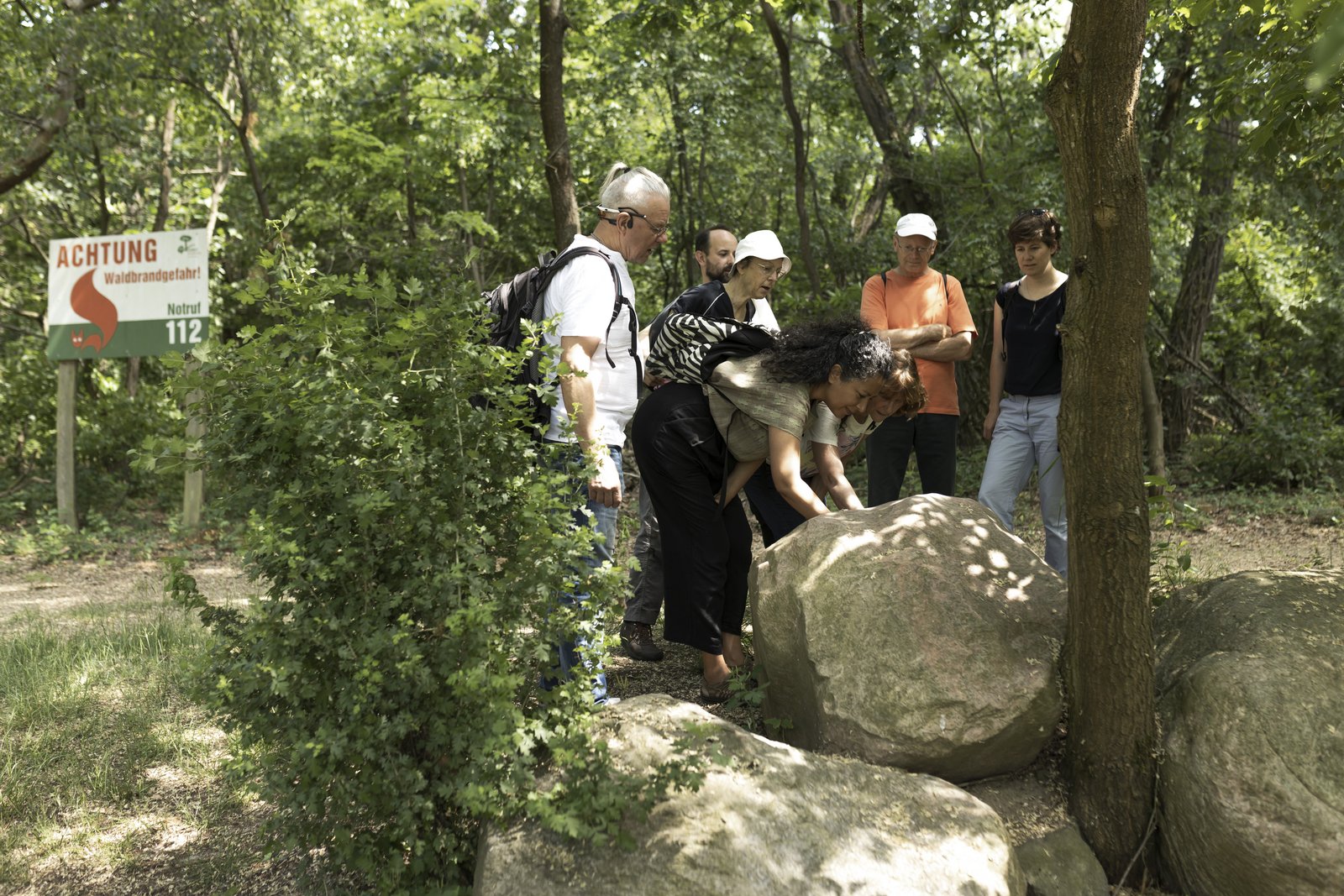 Fünf Personen stehen im Wald und schauen auf einen großen Stein. Eine Frau beugt sich über den Stein und bearbeitet die Oberfläche.  