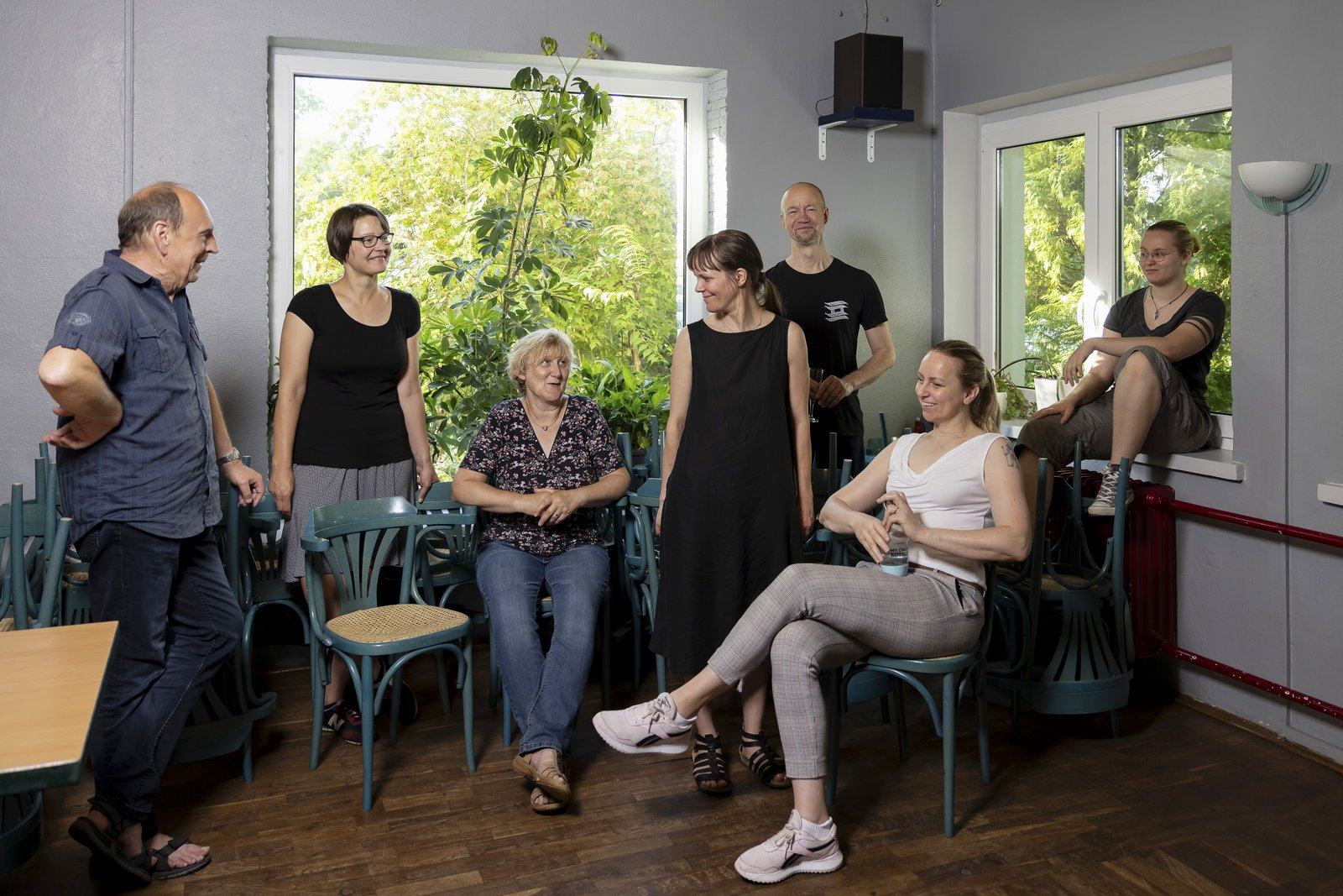 A group of seven people of different ages stand and sit in an interior room in front of open windows. They look and smile at each other. Green branches grow through the windows into the room.