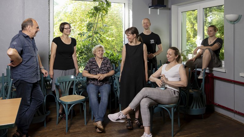 Eine Gruppe von sieben Personen unterschiedlichen Alters steht und sitzt in einem Innenraum vor geöffneten Fenstern. Sie schauen und lächeln sich gegenseitig an. Durch die Fenster ranken grüne Zweige in den Raum hinein.