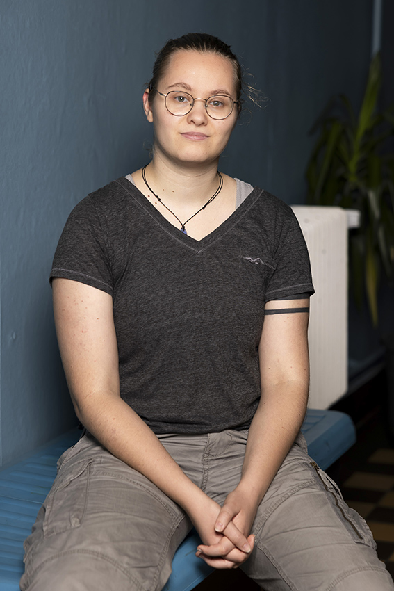 Eine Person mit Brille und blondem Zopf sitzt vor einer blauen Innenwand und schaut direkt in die Kamera.